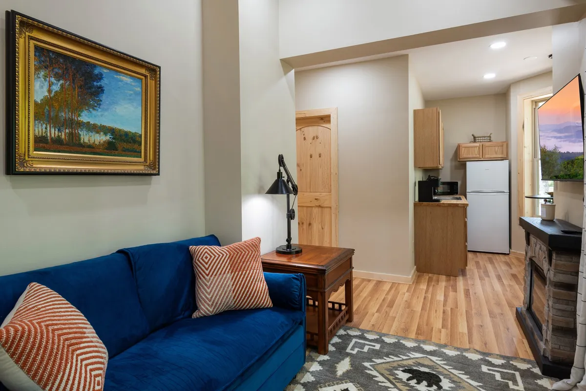 A cozy living area featuring a blue sofa, wooden furniture, and a kitchenette in the background. Stylish artwork and warm lighting help create an inviting, multifunctional space.