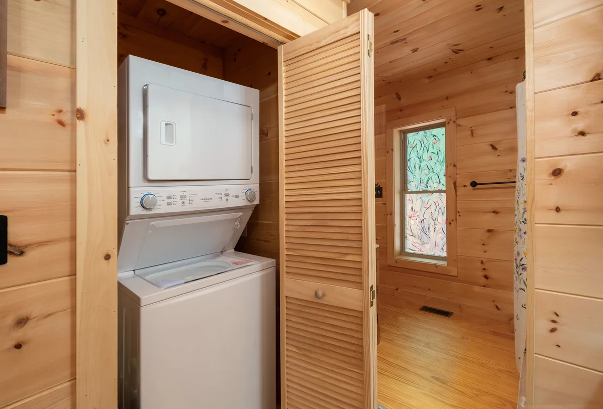 A tidy laundry closet featuring a washer-dryer unit behind a wooden slatted door, with a colorful window nearby. The space feels practical yet inviting, seamlessly integrated for guest convenience.