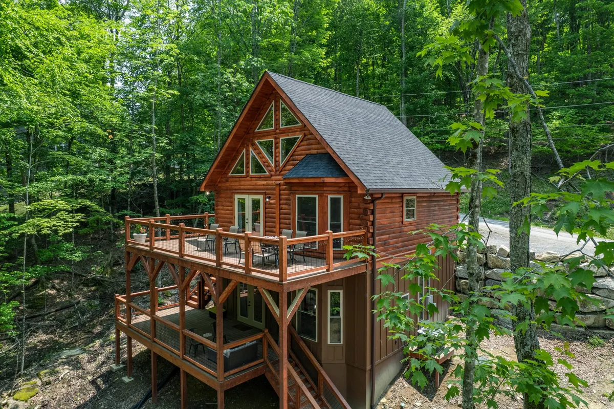 The image captures the striking exterior of a brand-new cabin nestled amongst tall trees on Beech Mountain. The centerpiece is a spacious wooden deck perfect for relaxing or gathering, complemented by large windows and a distinctive triangular roof that echoes the natural surroundings. The tranquil setting is intensified by the lush forest backdrop, inviting guests to immerse themselves in peaceful mountain living.