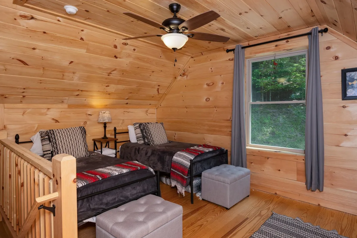 A cozy loft space with two twin beds beneath a wooden ceiling. A window invites in soft natural light and offers a view of the outdoors. The space is adorned with a decorative lamp and ceiling fan, making it perfect for children or additional guests.