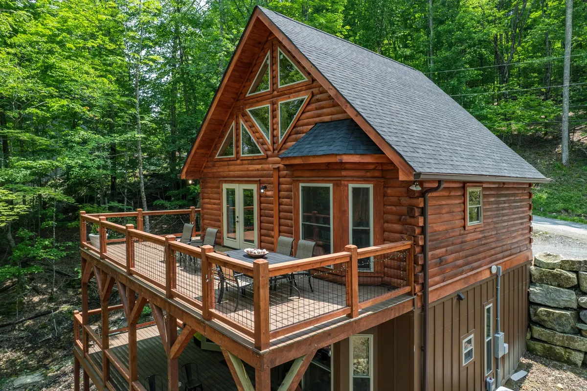 A charming log cabin with a spacious deck is nestled among lush trees. The triangular windows add a playful architectural touch, and the large deck invites outdoor relaxation, dining, or birdwatching. The setting feels welcoming for families and groups seeking connection with nature after exploring local attractions.