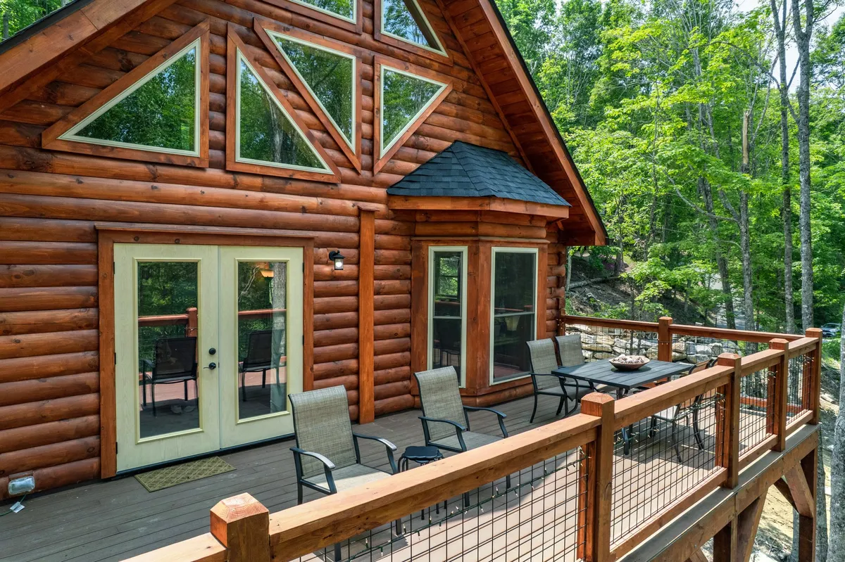 A calming exterior view of a log cabin with large windows and a spacious deck surrounded by towering trees. The deck is visible, inviting guests to relax and enjoy the sights and sounds of the forest, accentuated by the gentle glow of natural light.
