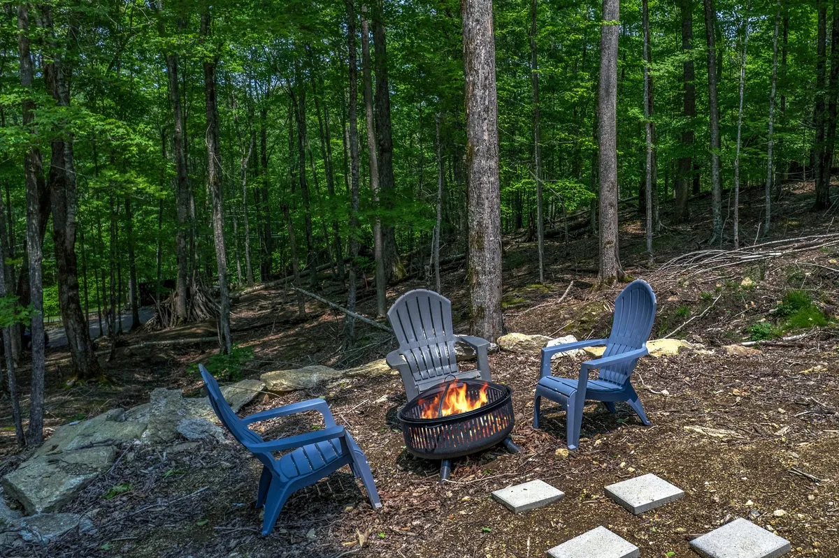 A cozy fire pit sits beneath a canopy of trees, encircled by classic Adirondack chairs. The area is ready for gatherings, marshmallow roasting, and storytelling, with the forest creating a natural, intimate enclosure.