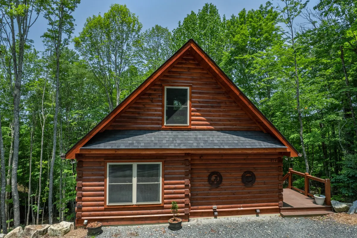 A log cabin with a large front window and a spacious wooden deck set amidst tall trees. The deck offers a cozy gathering space, perfect for outdoor relaxation and evening campfires. The setting evokes a sense of warmth, inviting guests to enjoy the outdoors as dusk falls.