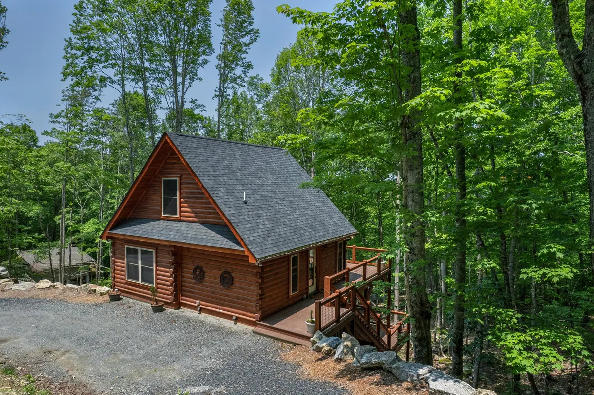This image features a wide exterior view of the log cabin, highlighting the spacious deck with railings and the surrounding wooded landscape. The gravel driveway is visible, emphasizing the importance of winter vehicle access, while the peaceful natural environment evokes a sense of mountain adventure and tranquility.