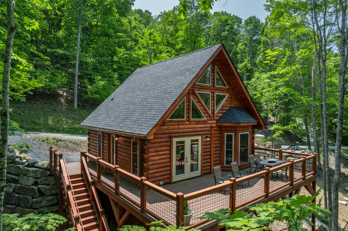 This image captures the log cabin's exterior, showcasing its inviting deck area nestled among tall trees. The scene radiates a cozy, peaceful mood, suggesting a warm retreat after a day of outdoor adventure. Subtle sunlight filters through the trees, illuminating the wooden structure and hinting at the beauty of mountain evenings.