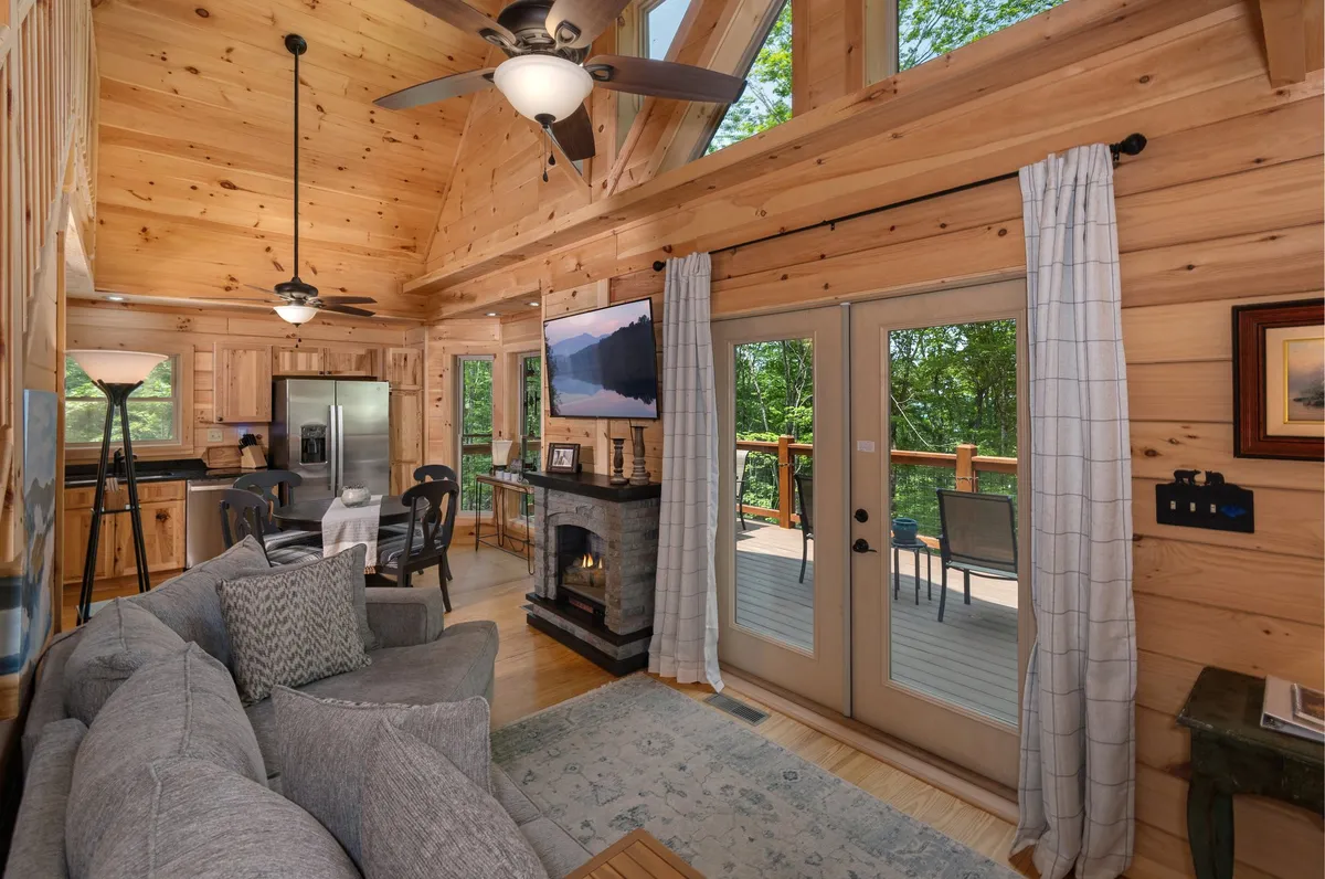 A wide-angle view of the open-concept main living area featuring a plush sofa, electric fireplace, expansive dining table, and large windows that flood the space with natural light. The room seamlessly connects the lounge, dining, and kitchen areas, with direct access to the deck and woodland views visible through the windows.