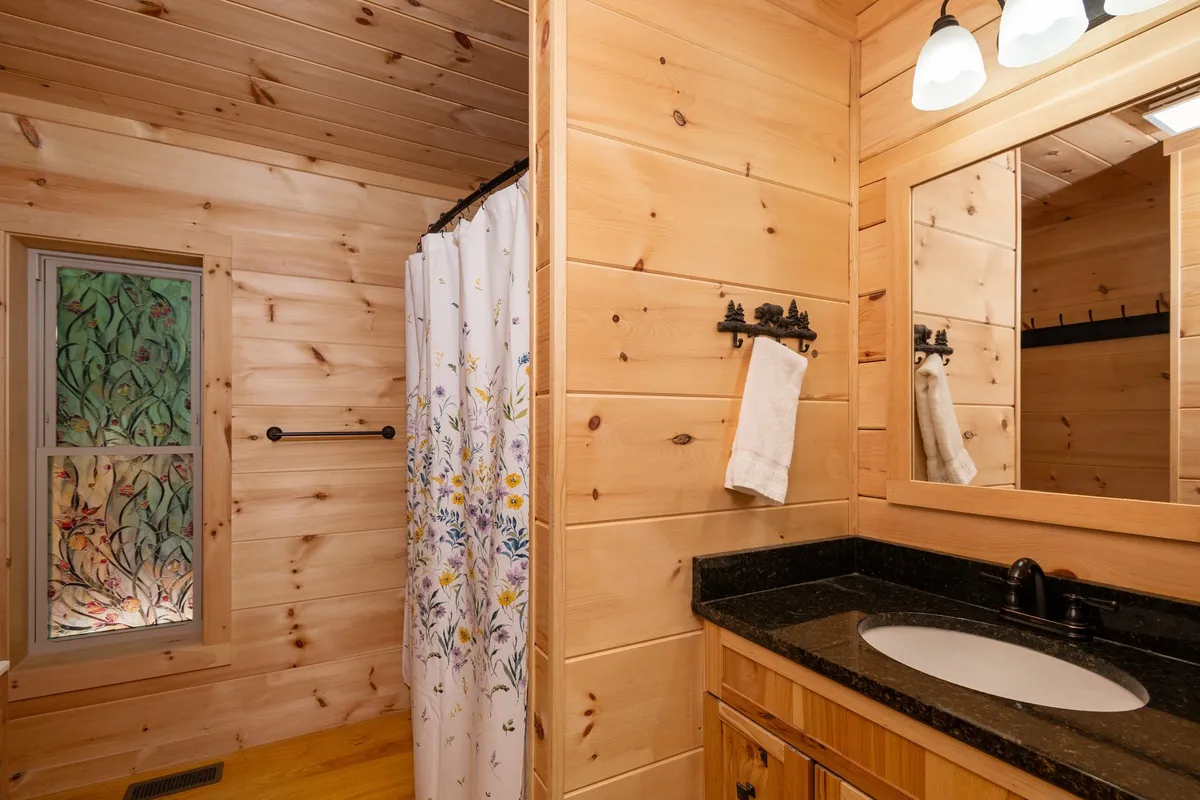 A full bathroom with wood paneling, a floral shower curtain, black granite countertop, and a well-lit mirror. The room is bathed in gentle natural light from a window, creating a calm and serene setting for self-care.