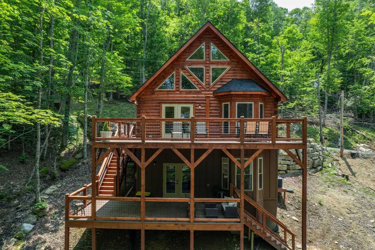 A tranquil wooden deck extends from the cabin, surrounded by tall trees and featuring large windows and a striking triangular roof. The deck is furnished for relaxation, with the forest canopy enveloping the space. The golden hues of a sunset hint at the perfect moments for morning coffee or evening wine outdoors.
